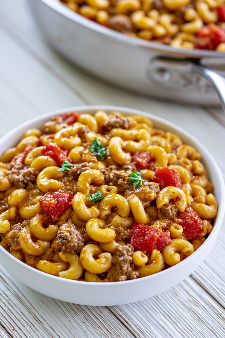 Bowl of taco mac and cheese made with elbow macaroni, seasoned ground beef, and diced tomatoes in a creamy orange cheese sauce, topped with a few parsley leaves, with the skillet blurred in the background.