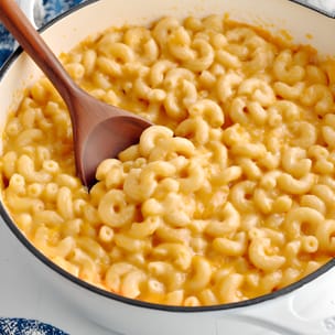 Creamy vegan mac and cheese in a white skillet, with elbow pasta coated in glossy orange cheese sauce and a wooden spoon scooping a serving.