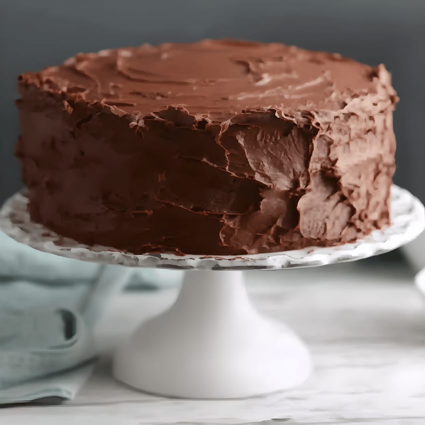 Frosted chocolate layer cake with thick, swirled chocolate buttercream on top and sides, shown on a white cake stand against a dark background.