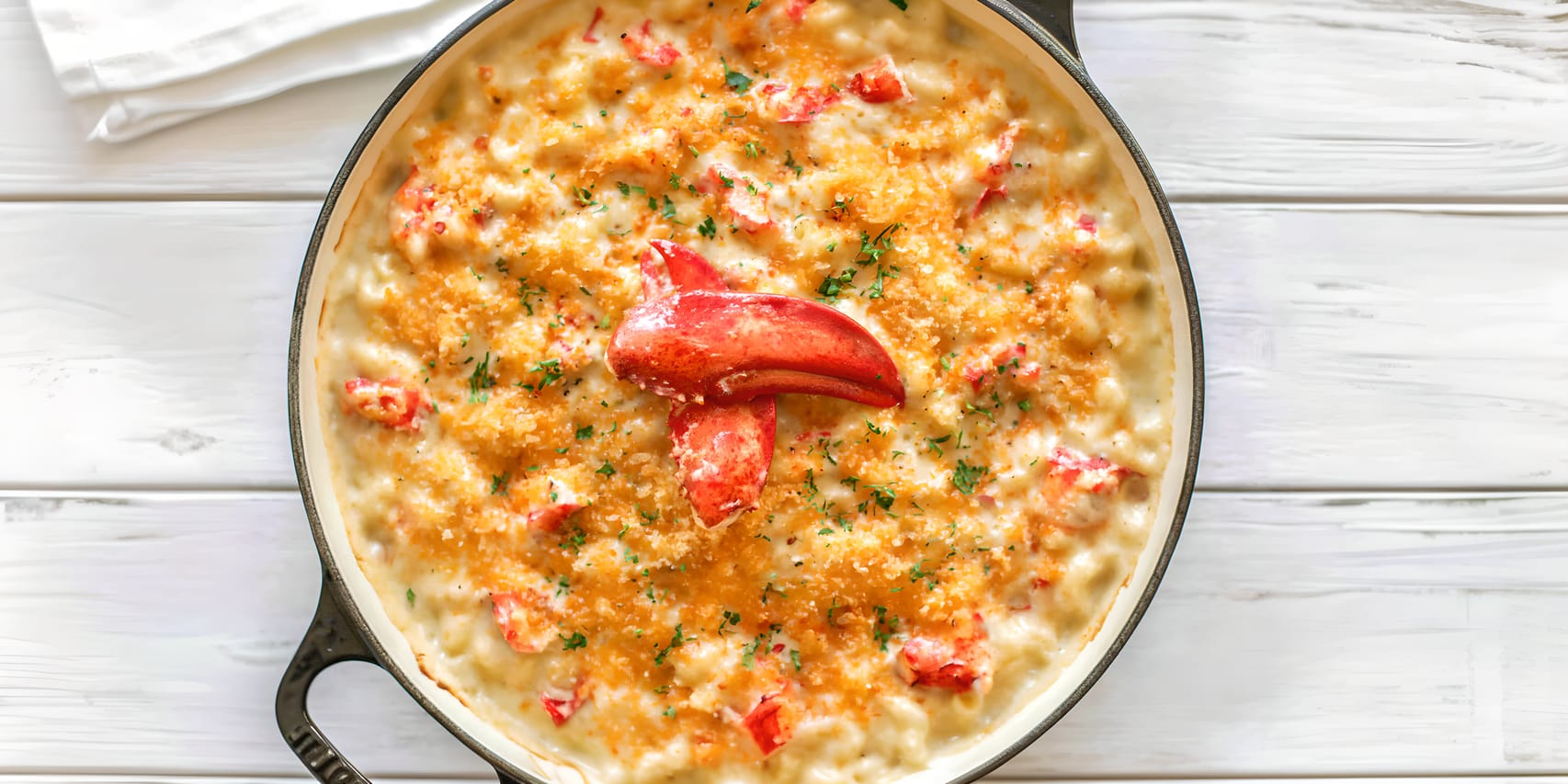 Overhead view of a skillet filled with creamy lobster mac and cheese, topped with golden toasted breadcrumbs, scattered lobster pieces, chopped herbs, and a large red lobster claw in the center.