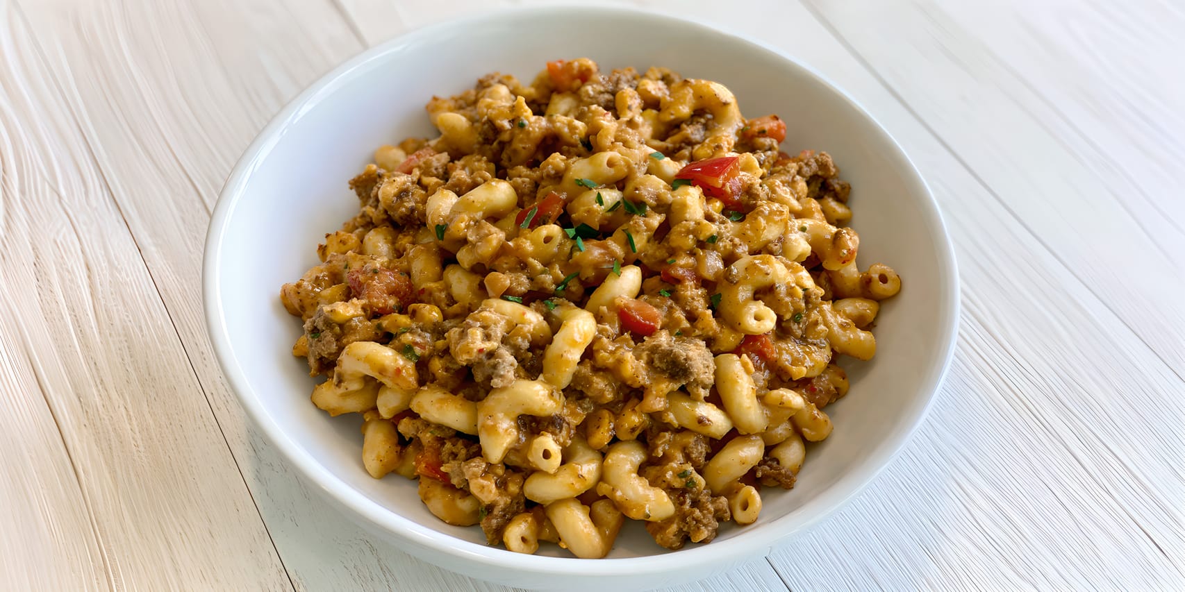 Bowl of taco mac and cheese made with elbow macaroni, browned ground beef, and a creamy orange cheese sauce, with visible diced tomatoes and green herb garnish, served in a white bowl on a light wood surface.