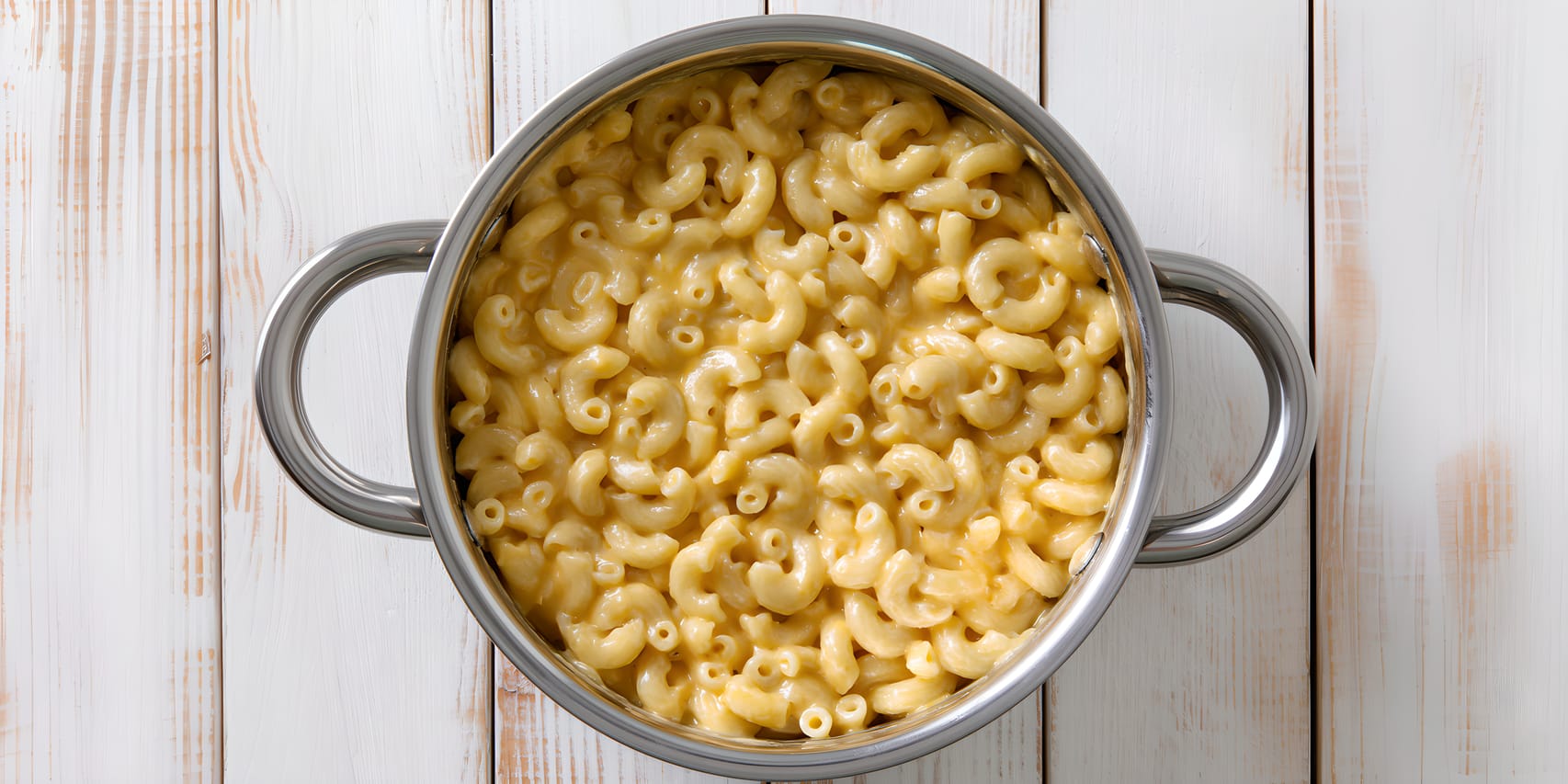 Overhead view of creamy macaroni and cheese in a stainless steel pot with side handles, set on a whitewashed wooden surface.