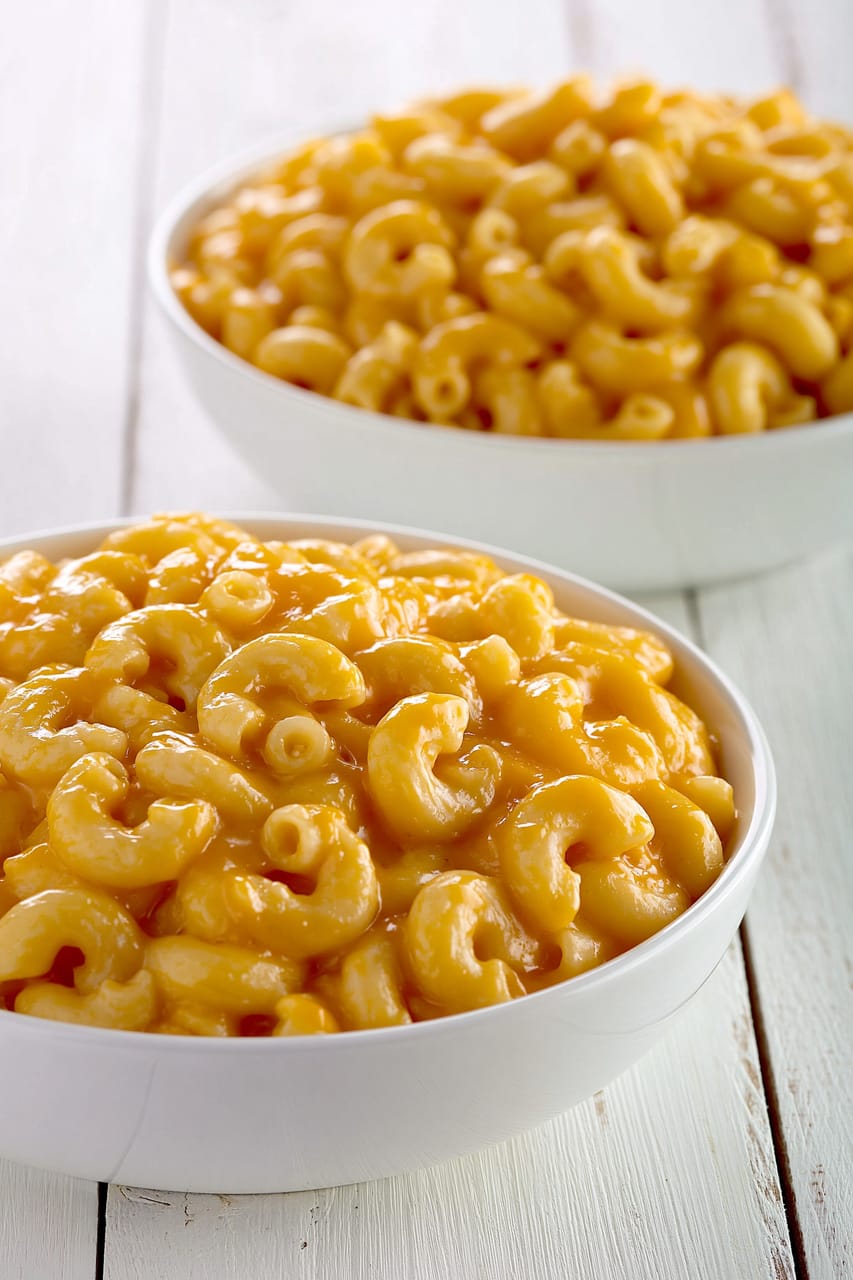Two white bowls of elbow macaroni coated in a thick, glossy orange nacho cheese sauce on a white wooden surface, with the front bowl in sharp focus and the back bowl softly blurred.