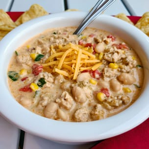 White bowl of creamy ground-chicken chili topped with shredded cheddar, with visible white beans, corn, diced tomatoes, and green chiles; a spoon rests in the bowl and tortilla chips surround it on a red napkin.