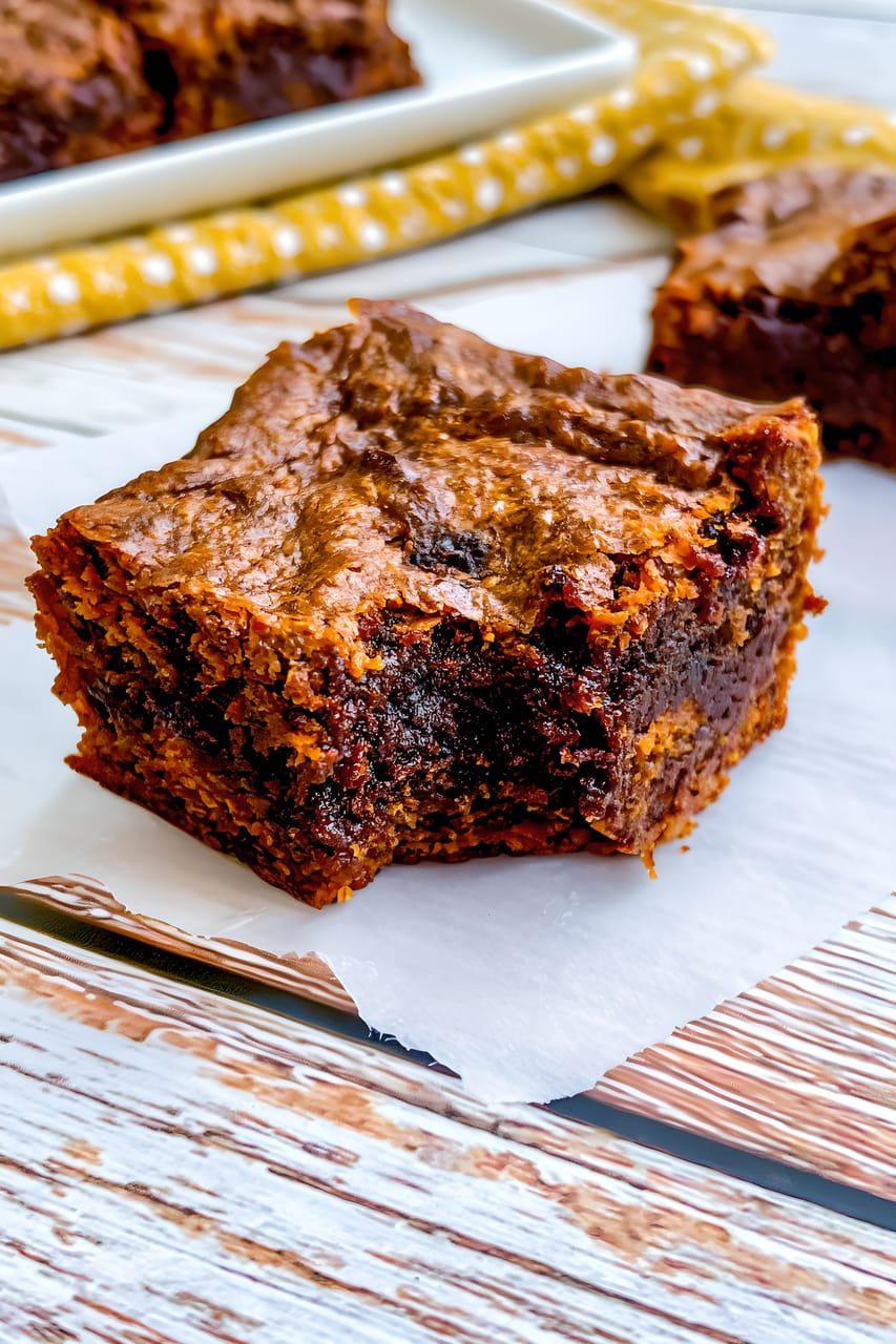 Close-up of a thick square sweet potato brownie on a small sheet of parchment, showing a shiny crinkled top and dense, fudgy chocolate center. More brownie pieces sit softly blurred in the background near a white baking dish and yellow patterned cloth.