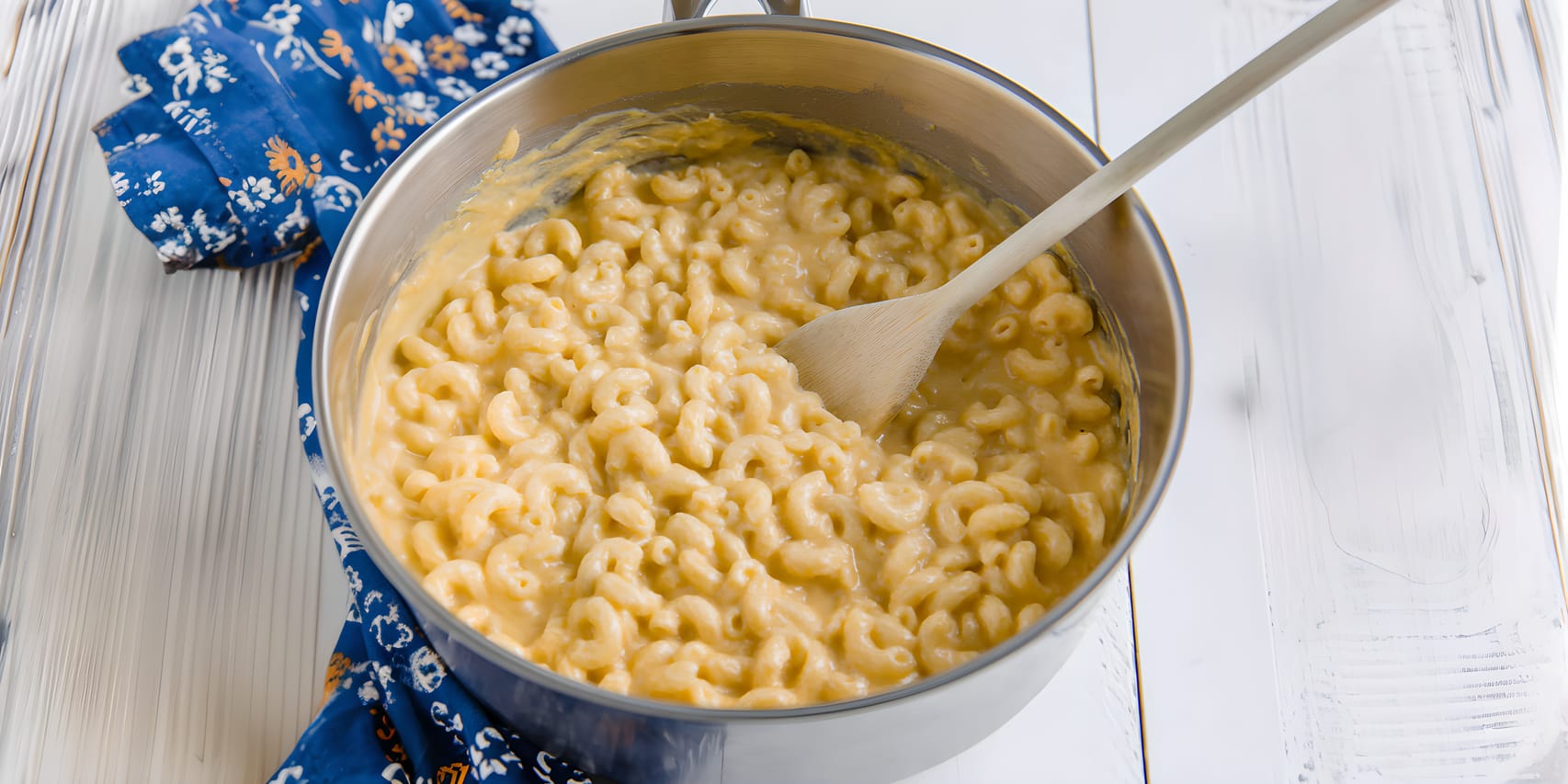 Creamy high-protein mac and cheese in a large metal pot, with elbow macaroni coated in a thick pale cheddar sauce and a wooden spoon resting inside. The pot sits on a white wood surface with a blue patterned cloth underneath.
