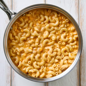 Overhead view of a stainless steel saucepan filled with creamy spicy nacho cheese mac and cheese. Elbow macaroni is coated in a smooth orange cheese sauce with small flecks of red pepper, shown on a white wooden surface.