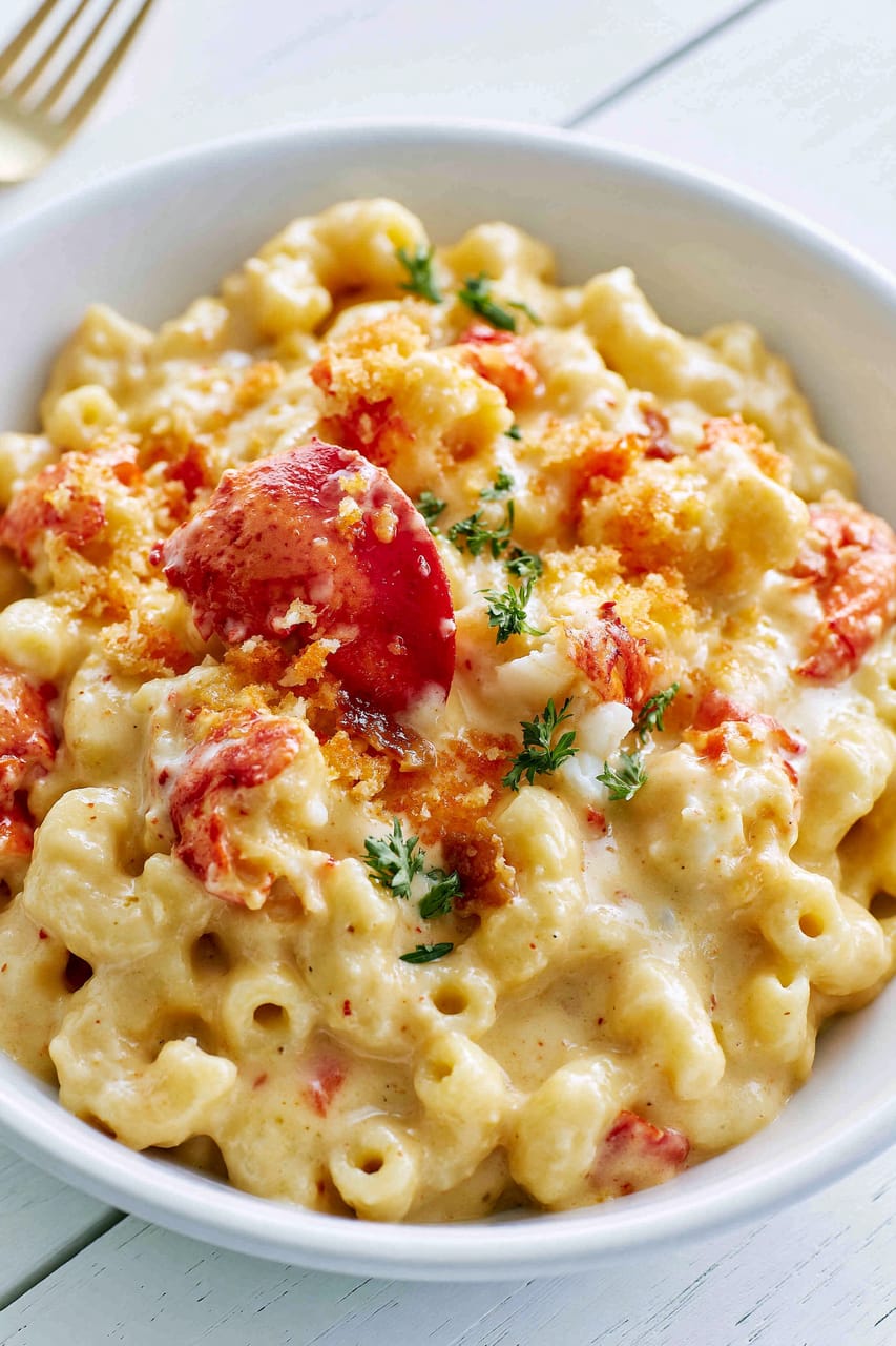Close-up of a white bowl filled with creamy lobster mac and cheese, with elbow pasta coated in a thick pale-gold cheese sauce, chunks of red lobster, toasted golden breadcrumbs, and a sprinkle of green herbs on top.