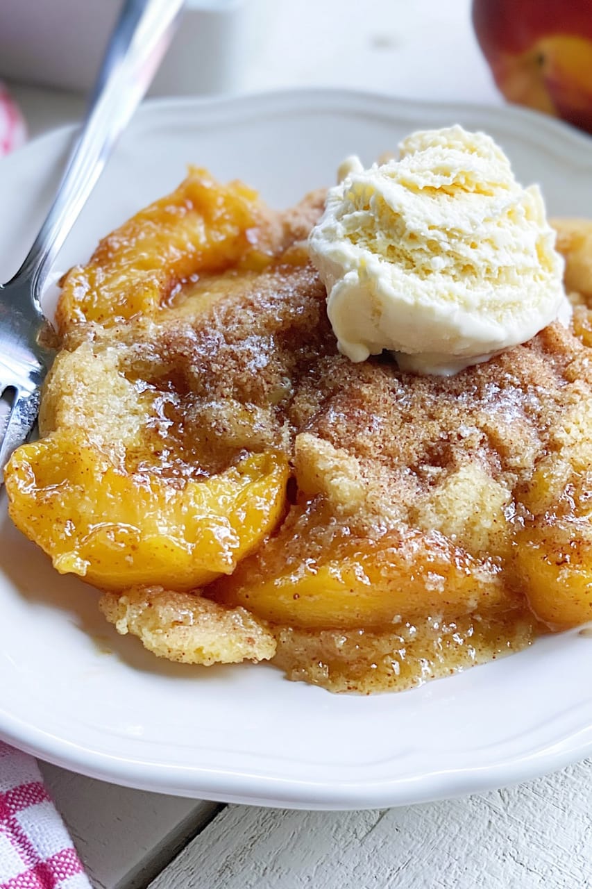 Serving of peach cobbler on a white plate, with glossy cinnamon-spiced peach slices and a golden, cake-like topping, finished with a scoop of vanilla ice cream melting over the top.
