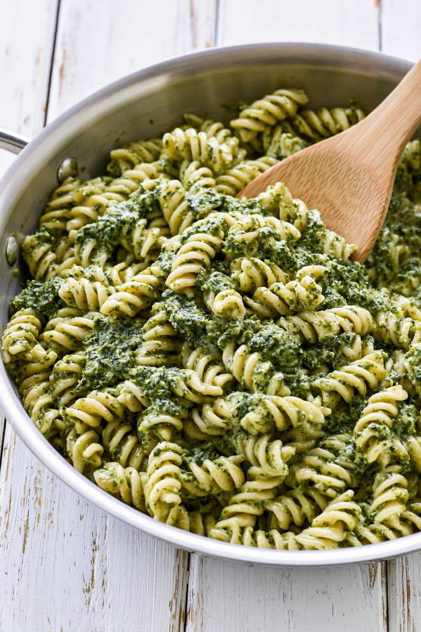 Close-up of rotini pasta in a stainless steel skillet, coated in a creamy green spinach sauce with visible flecks of spinach. A wooden spoon is stirring the pasta, and the pan sits on a white wooden surface.