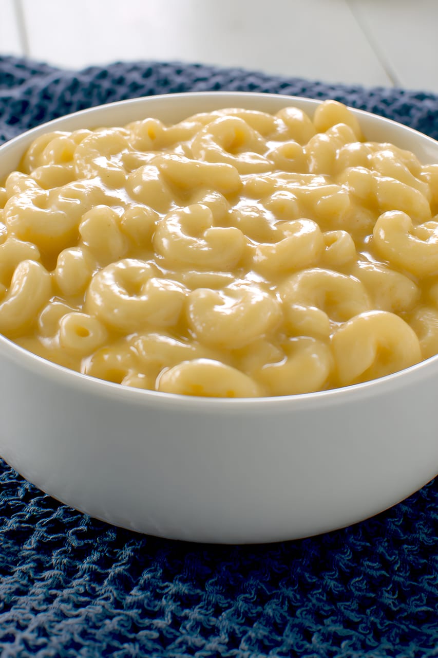 Close-up of a white bowl filled with creamy elbow macaroni and cheese, with a smooth, glossy pale yellow sauce, set on a dark blue textured cloth.