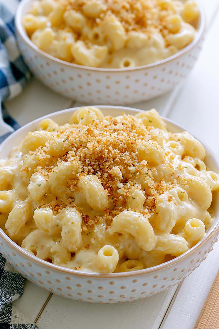 Two white dotted bowls of creamy Spanish Manchego mac and cheese, topped with golden toasted panko breadcrumbs. The front bowl is in sharp focus, showing glossy elbow pasta coated in thick cheese sauce, with a second bowl softly blurred in the background on a light table.