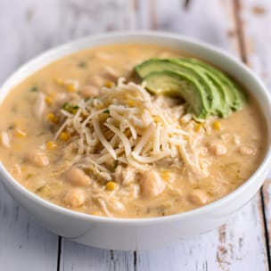 Bowl of creamy white chicken chili topped with shredded cheese and sliced avocado, with visible shredded chicken, cannellini beans, and corn in a pale golden broth.