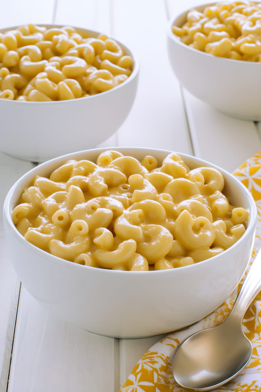 Three white bowls of creamy macaroni and cheese on a white wooden table, with the front bowl in focus showing glossy elbow pasta coated in smooth cheddar sauce. A silver spoon and yellow patterned napkin sit at the lower right.