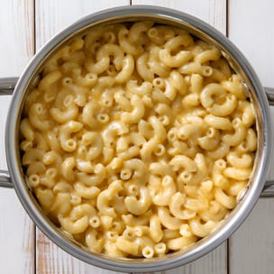 Overhead view of a stainless steel pot filled with creamy elbow macaroni and cheese, the noodles evenly coated in a smooth, glossy cheddar sauce on a white wooden surface.