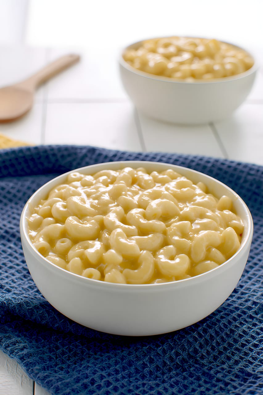 Close-up of creamy macaroni and cheese in a white bowl, with glossy elbow pasta coated in a smooth cheese sauce. The bowl sits on a dark blue textured towel, with a second blurred bowl and a wooden spoon in the bright background.