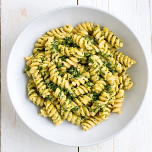 Overhead view of a white bowl filled with rotini pasta coated in a creamy pale-yellow sauce with finely chopped spinach throughout, set on a white wooden surface.