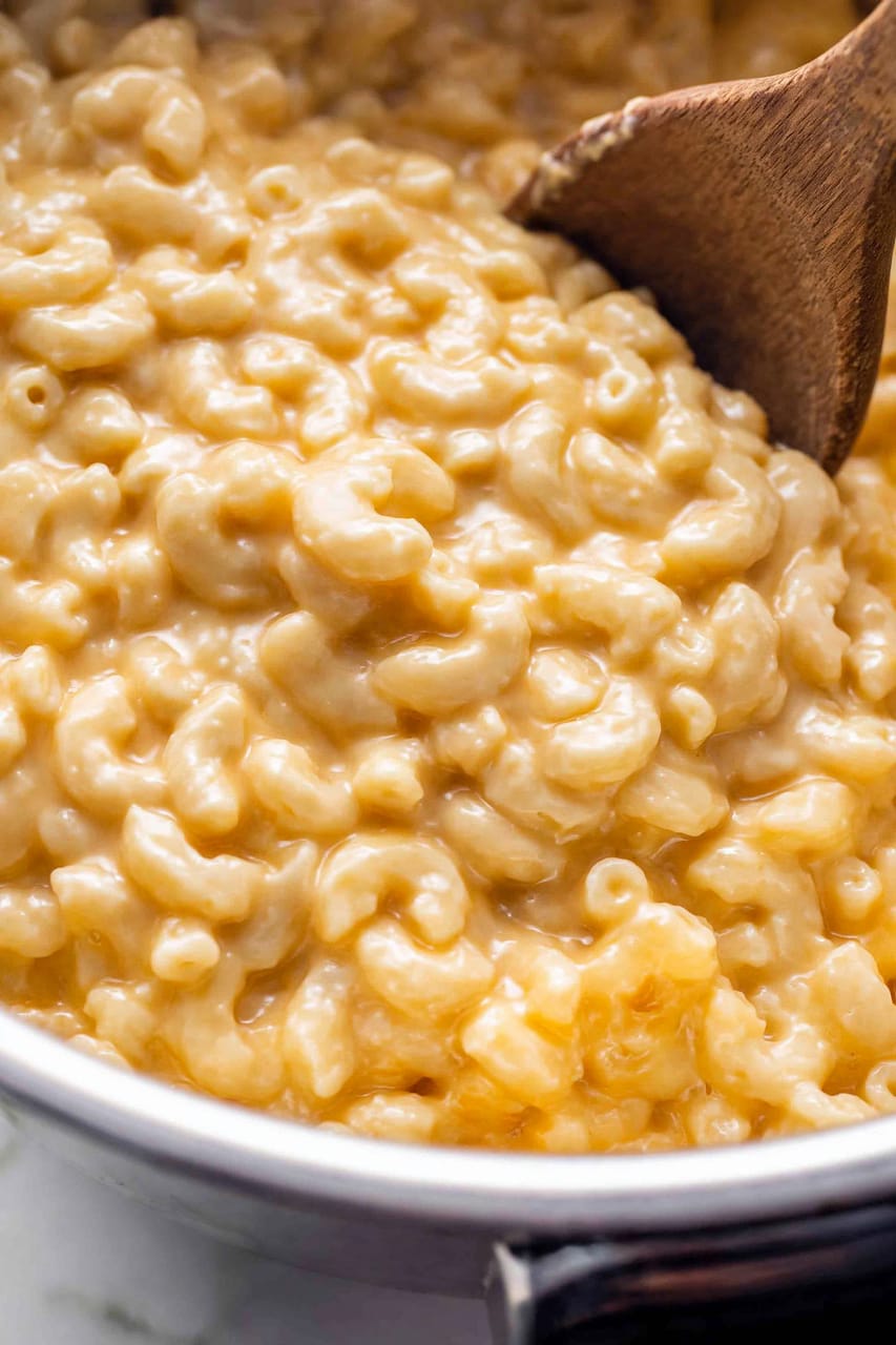 Close-up of creamy keto macaroni and cheese in a pot, with elbow-shaped pasta coated in thick, glossy orange cheese sauce and a wooden spoon stirring through it.