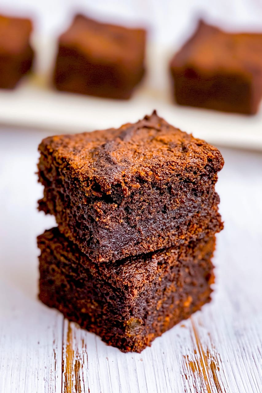 Close-up of two stacked sweet potato brownies on a whitewashed surface, showing a crackly top and dense, fudgy chocolate center, with more brownie squares blurred in the background.