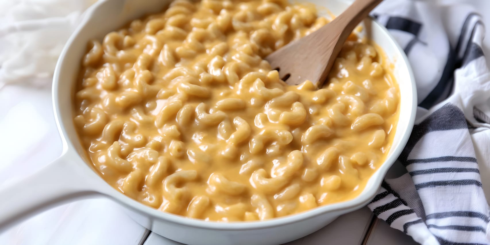 Creamy Velveeta mac and cheese in a white serving dish, with elbow macaroni coated in a smooth, glossy orange cheese sauce and a wooden spoon resting in the dish beside a striped kitchen towel.