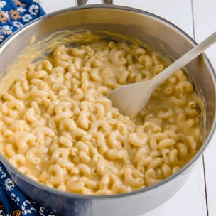 Creamy elbow macaroni and cheese in a stainless steel pot, stirred with a wooden spoon; the glossy cheese sauce thickly coats the pasta, showing the finished stovetop dish.
