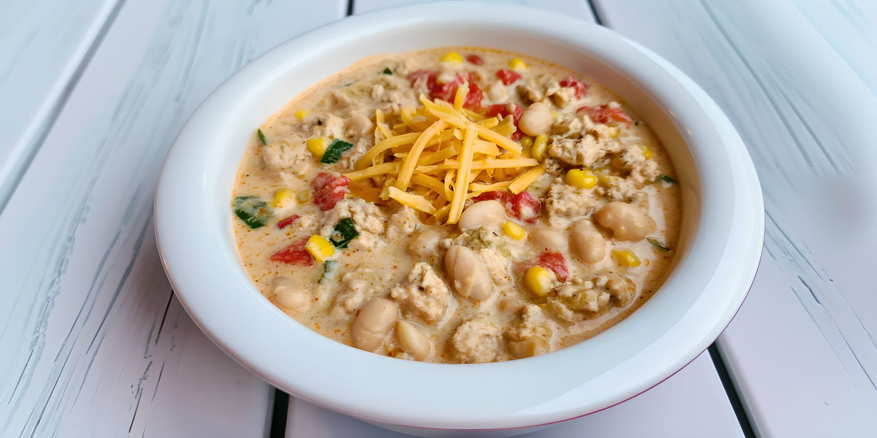 Bowl of creamy white ground-chicken chili topped with shredded cheese, with visible Great Northern beans, corn, and diced tomatoes in the rich broth.