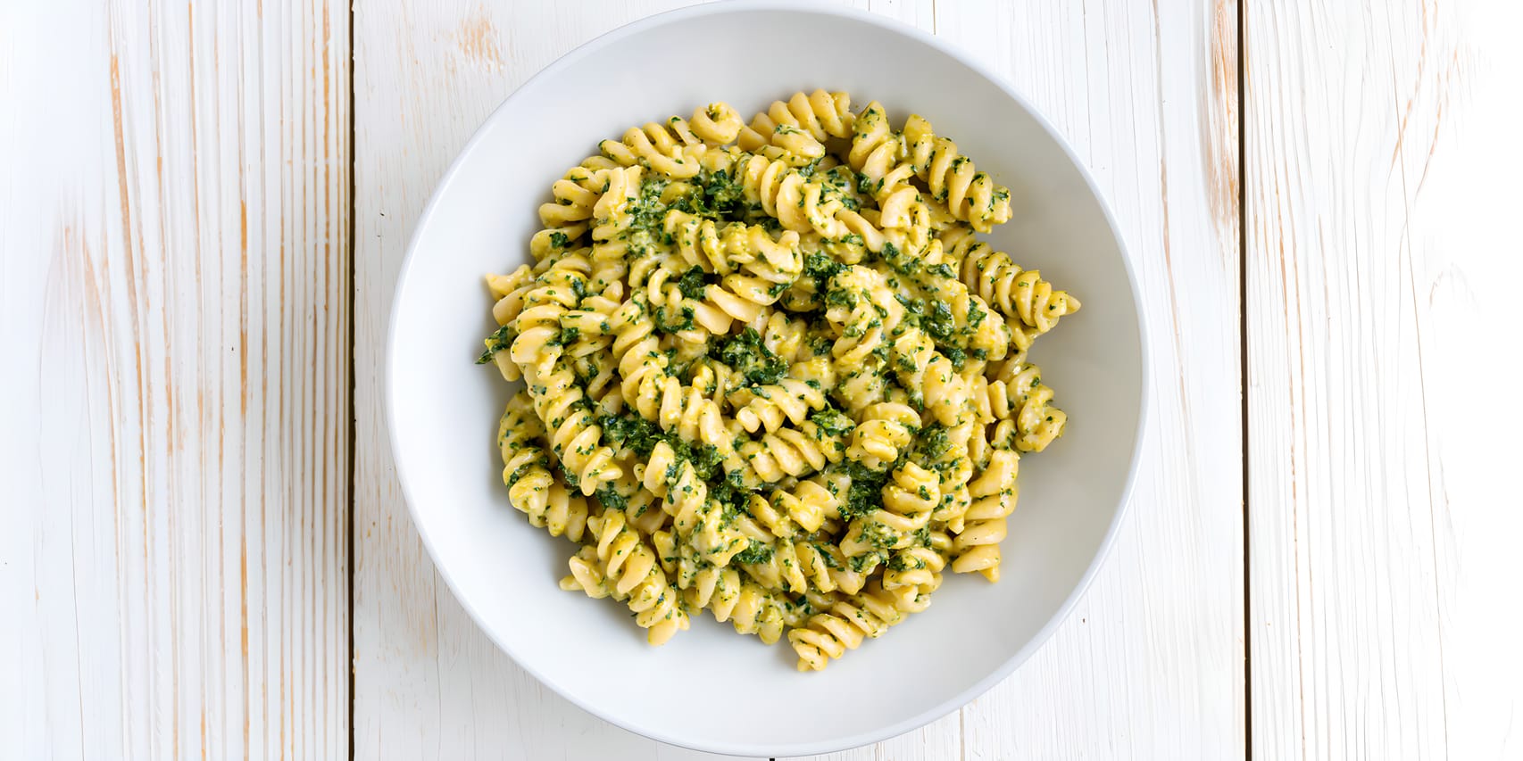 Overhead view of a white bowl filled with rotini pasta coated in a creamy pale-yellow sauce with finely chopped spinach, set on a white wooden surface.