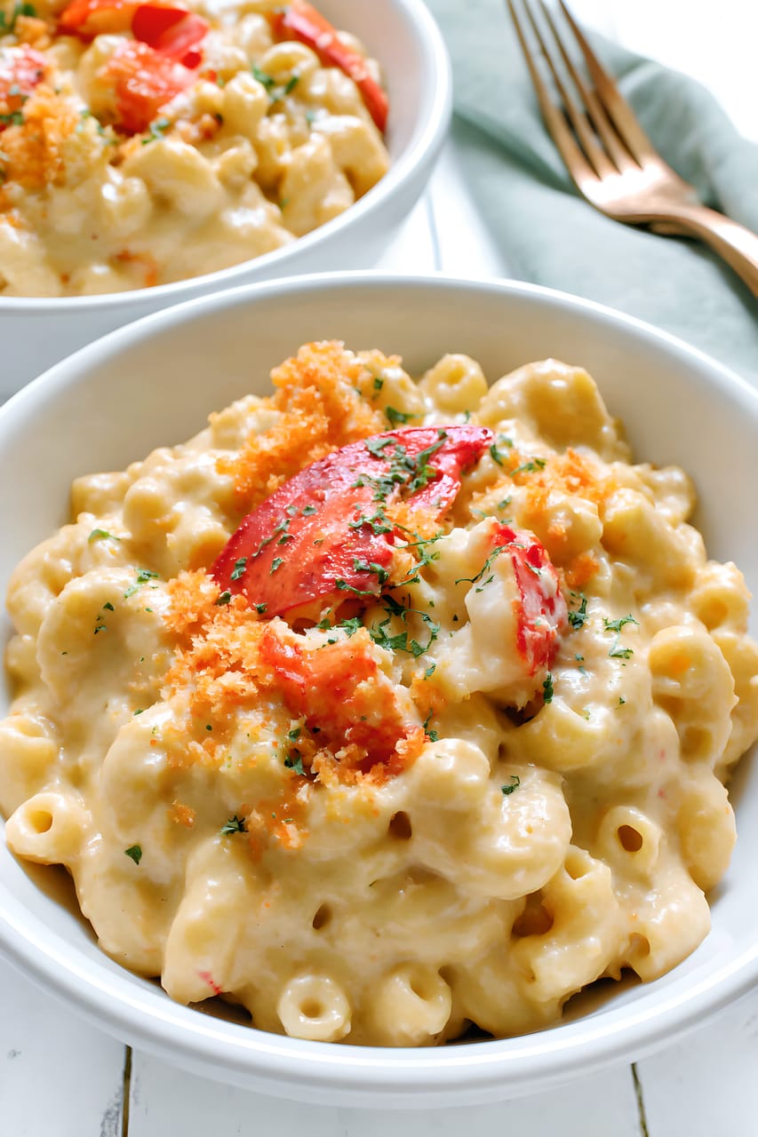 Two white bowls of lobster mac and cheese with creamy cavatappi pasta, topped with red lobster pieces, golden toasted breadcrumbs, and chopped chives; a fork and pale green napkin sit beside the bowls.