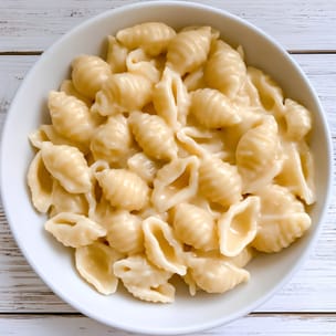 Overhead view of white cheddar mac and cheese made with shell pasta, coated in a thick, glossy pale cream-colored cheese sauce, served in a white bowl on a white wooden surface.