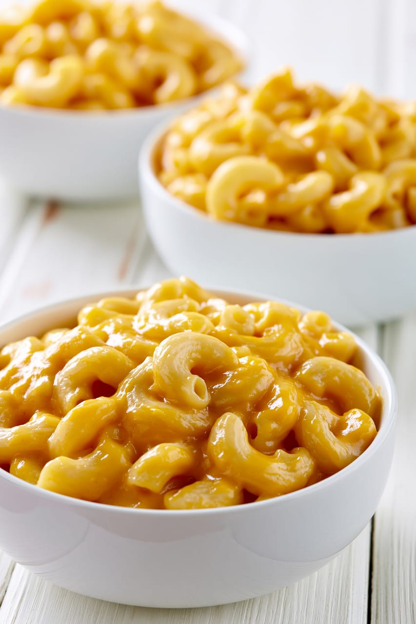 Three white bowls of creamy Velveeta mac and cheese on a light wooden surface, with the front bowl in sharp focus showing elbow macaroni coated in a thick, glossy orange cheese sauce.