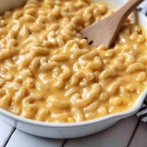 Close-up of creamy Velveeta mac and cheese in a white baking dish, with elbow macaroni coated in a smooth, glossy orange cheese sauce and a wooden spoon stirring through it.