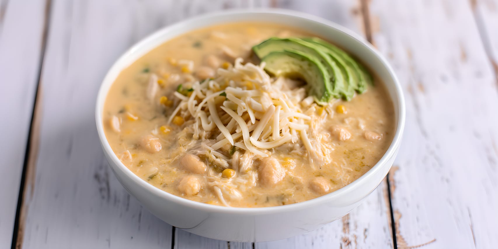 Bowl of creamy white chicken chili topped with shredded cheese and sliced avocado, with visible white beans, corn, and shredded chicken in the pale golden broth.