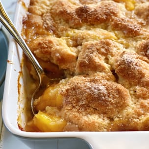 Close-up of baked peach cobbler in a white casserole dish, with a spoon scooping out glossy peach filling beneath a golden-brown biscuit-style topping dusted with sugar.