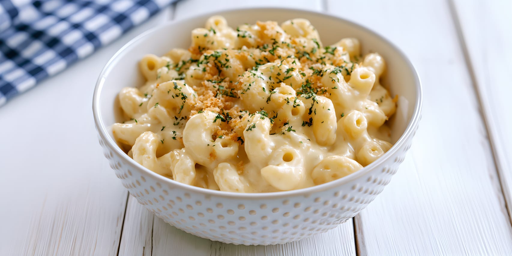 Bowl of creamy Spanish Manchego mac and cheese on a white wooden table, with pasta coated in a glossy pale cheese sauce and topped with golden toasted breadcrumbs and a sprinkle of chopped parsley.