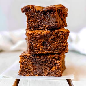 Three sweet potato brownies stacked on parchment, showing a dark chocolate color, thin crackly tops, and a dense, fudgy interior with melted chocolate throughout.