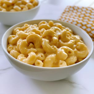 Bowl of creamy homemade macaroni and cheese, with elbow pasta coated in a thick, glossy cheddar sauce. A second bowl sits blurred in the background beside a yellow-and-white checkered napkin on a light countertop.