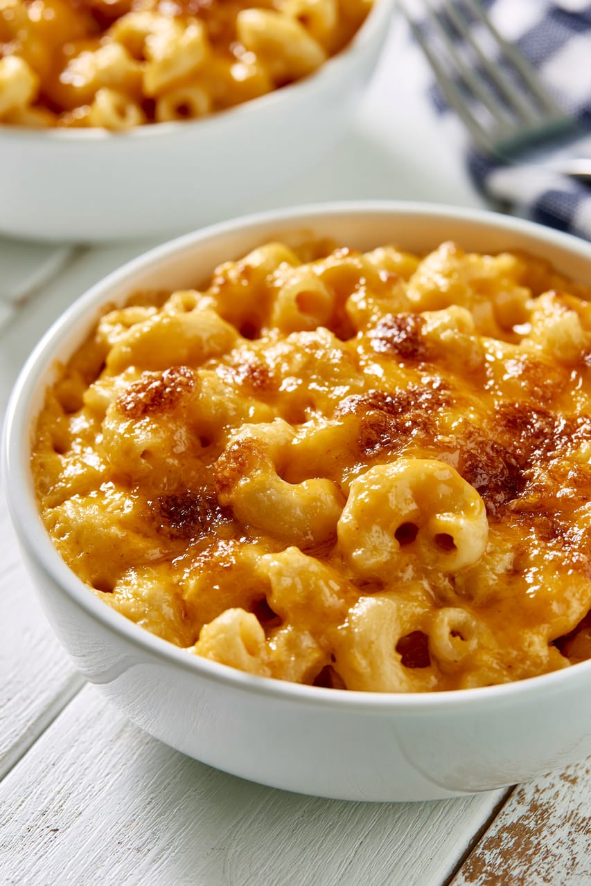 Close-up of baked Southern mac and cheese in a white bowl, with elbow macaroni in a creamy orange cheese sauce and a browned, bubbly top. A larger serving dish is softly blurred in the background on a white wooden table.