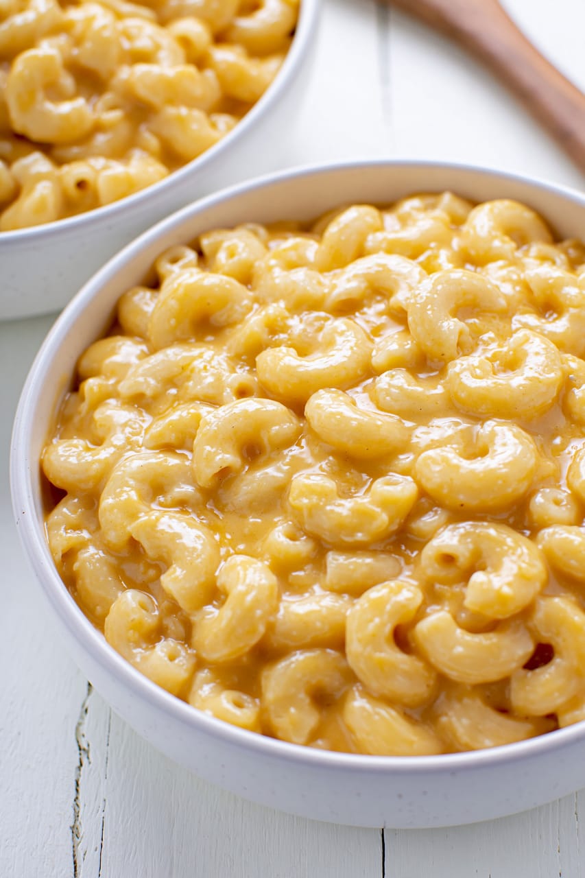 Two white bowls of keto mac and cheese filled with elbow pasta coated in a thick, glossy orange cheese sauce, shown in a close-up on a white wooden surface.