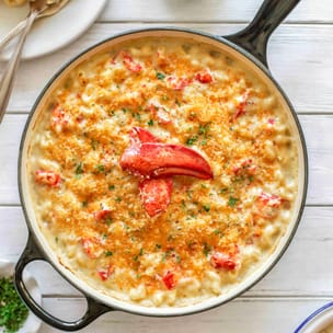 Overhead view of lobster mac and cheese in a white enameled skillet, with creamy cavatappi in a rich cheese sauce topped with golden toasted breadcrumbs, scattered lobster pieces, chopped herbs, and a bright red lobster claw in the center.