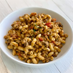 Bowl of taco mac and cheese made with elbow macaroni, seasoned ground beef, and red tomato pieces in a creamy orange cheese sauce, lightly sprinkled with chopped herbs.