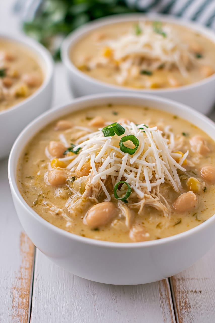Three white bowls of creamy white chicken chili on a rustic white table; the front bowl is topped with shredded white cheese and sliced green onions, with visible cannellini beans, corn, and shredded chicken in the pale golden broth.