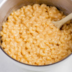 Close-up of creamy keto macaroni and cheese in a pot, with elbow pasta coated in a thick orange cheese sauce and a wooden spoon stirring from the side.