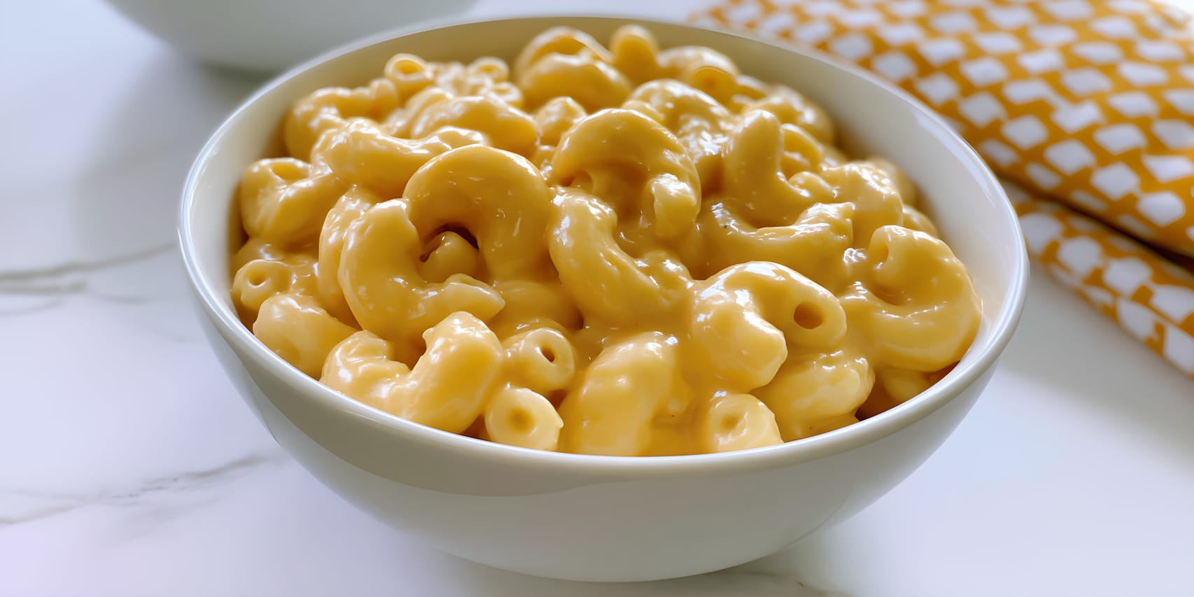 Bowl of creamy homemade mac and cheese, with elbow macaroni coated in a thick, glossy golden cheese sauce, served on a white surface beside a yellow patterned napkin.