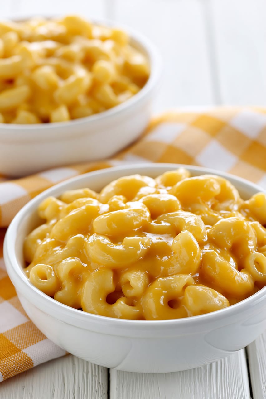 Close-up of creamy Velveeta mac and cheese in a white bowl, with glossy elbow macaroni coated in thick orange cheese sauce. A second serving dish sits softly blurred in the background on a yellow-and-white checkered cloth.