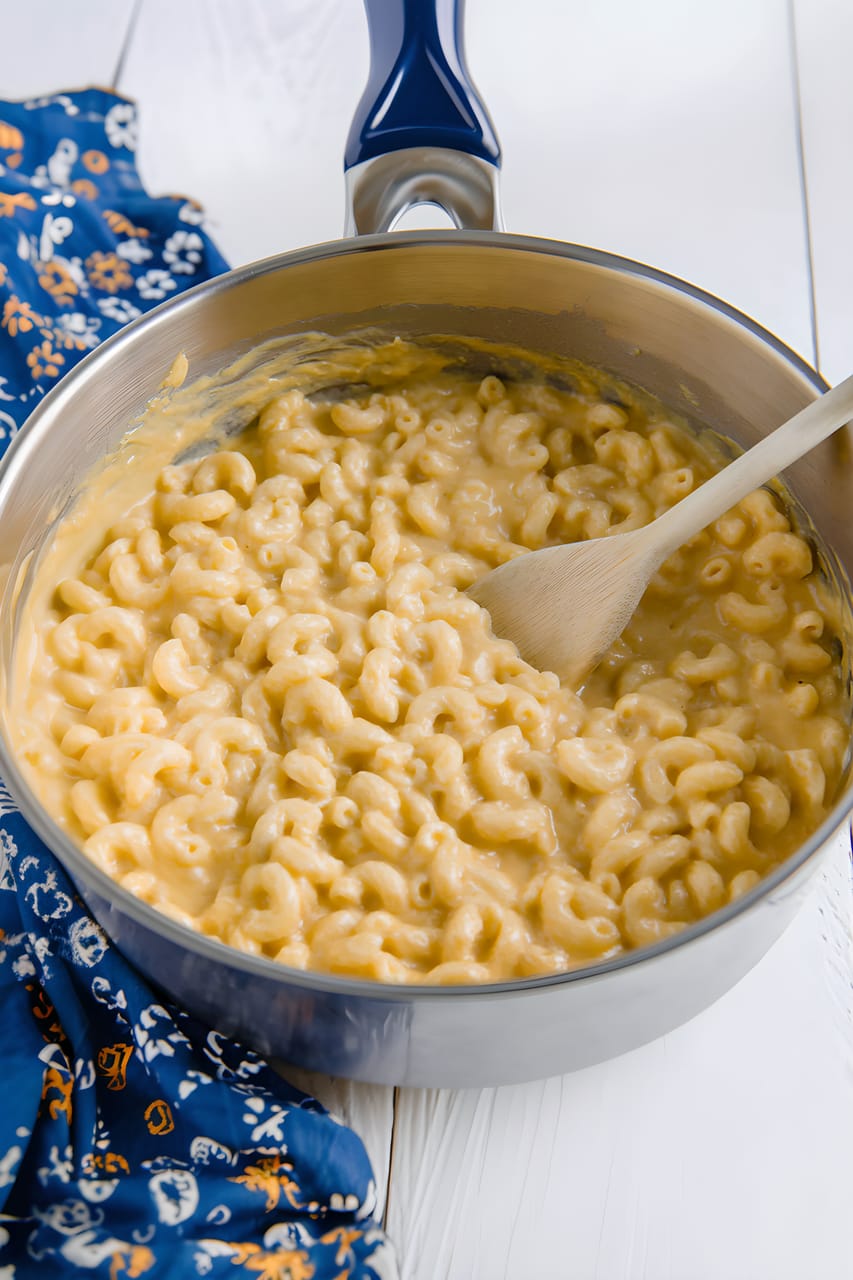 Creamy elbow macaroni and cheese in a stainless steel saucepan, with a wooden spoon resting in the glossy orange cheese sauce on a white tabletop.