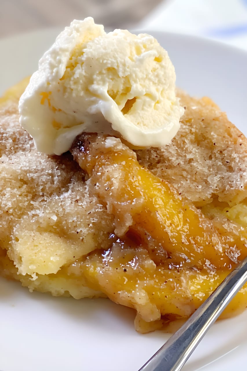 Close-up of a serving of peach cobbler on a white plate, with golden biscuit-like topping over glossy cinnamon-speckled peaches and a scoop of vanilla ice cream melting on top; spoon visible at the edge.