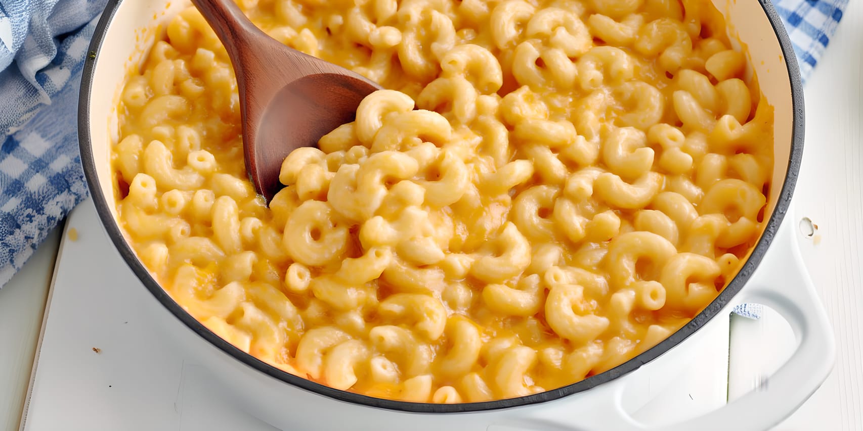 Close-up of creamy vegan mac and cheese in a white pot, with elbow pasta coated in a glossy orange sauce and a wooden spoon scooping from the center; blue check cloths sit in the background.