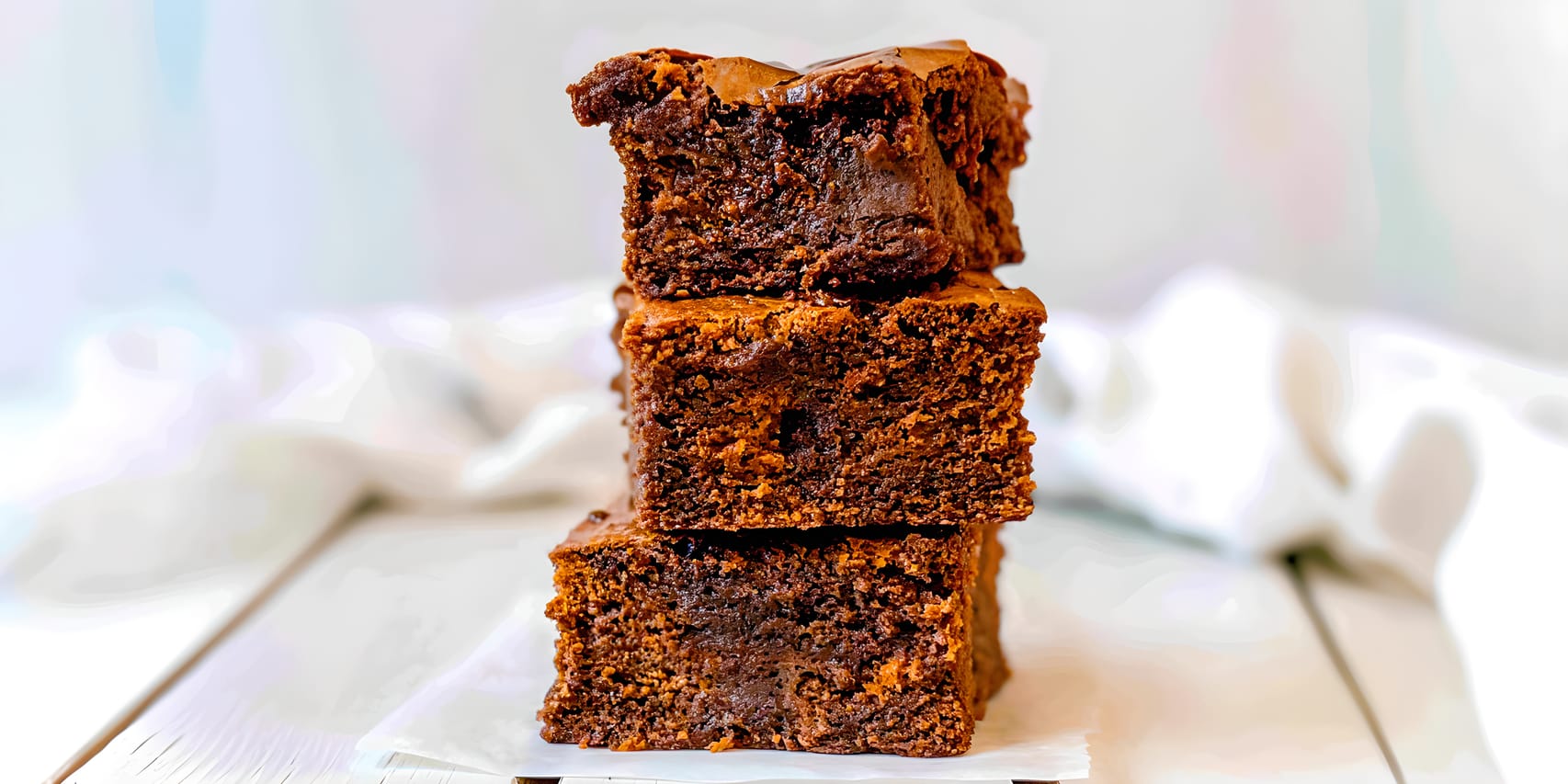 Three thick sweet potato brownies stacked on parchment paper, showing a dark, fudgy center with a thin crackly top and visible chocolate throughout, against a softly blurred light background.