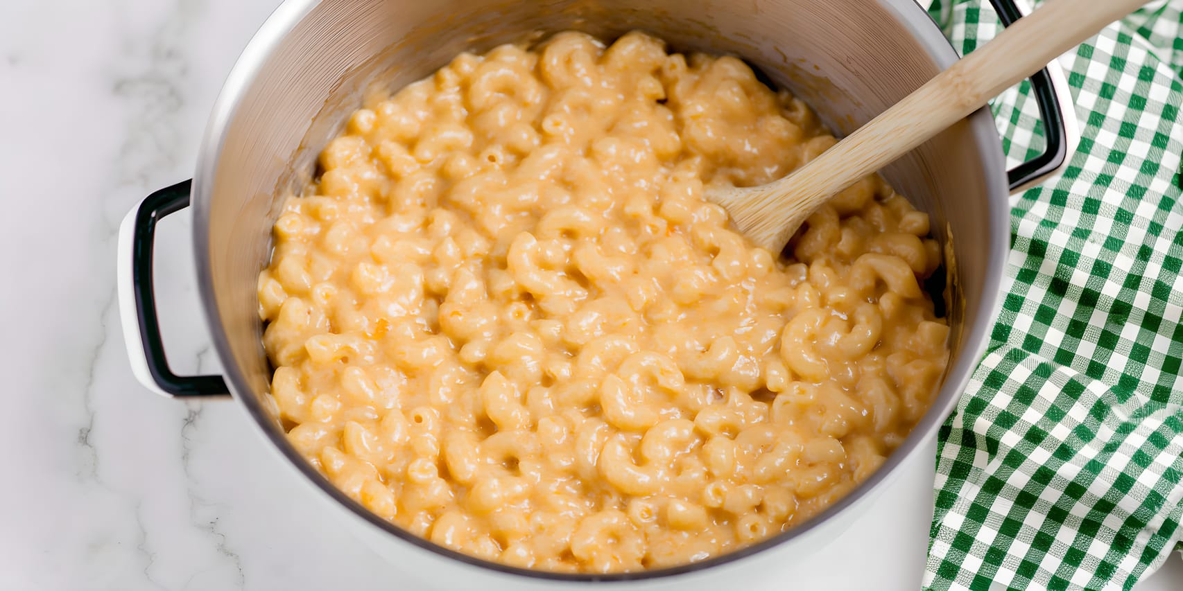 Creamy keto mac and cheese in a large pot, with elbow-shaped pasta coated in a thick orange cheese sauce and a wooden spoon resting inside; a green checkered kitchen towel sits beside the pot.