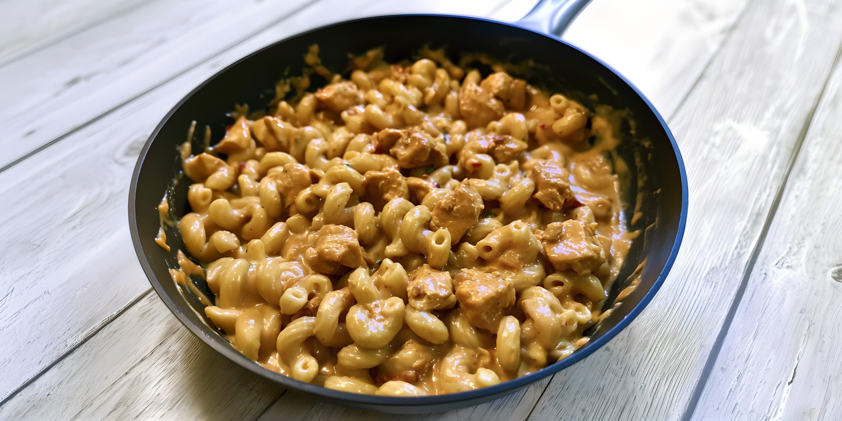 Skillet filled with creamy chipotle chicken mac and cheese, showing elbow macaroni and bite-size chicken pieces coated in a glossy orange cheese sauce on a whitewashed wooden surface.